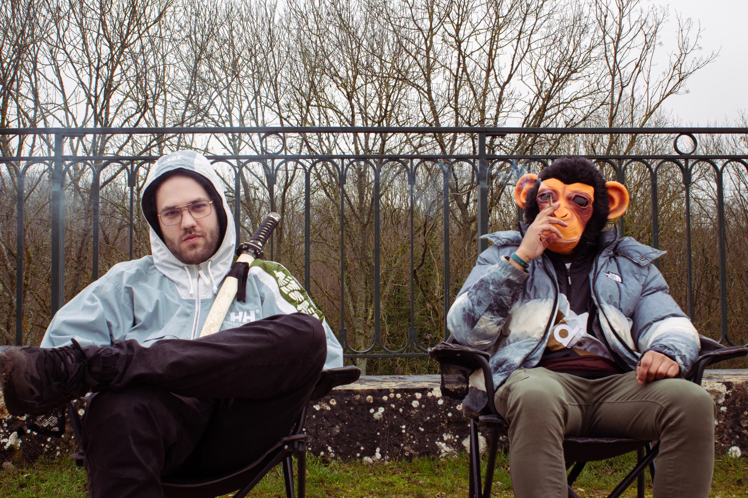 Deux hommes posent assis, l’un avec une batte, l’autre masqué en singe, ambiance insolite.
